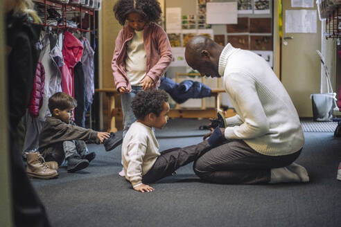 Side view of male teacher helping boy while wearing shoe at kindergarten - MASF43124