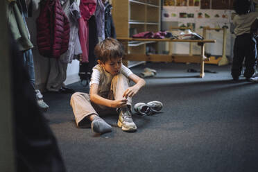 Boy tying shoelace while sitting on floor in cloakroom at preschool - MASF43125