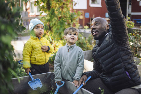 Male teacher talking with preschool kids holding shovel at garden - MASF43130