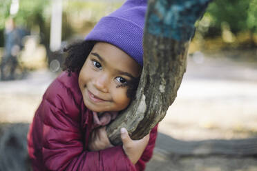 Portrait of smiling girl wearing knit hat and embracing tree branch at park - MASF43134
