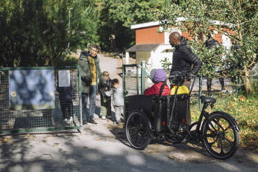 Female teacher standing at gate while welcoming children sitting in cargo bike at kindergarten - MASF43135