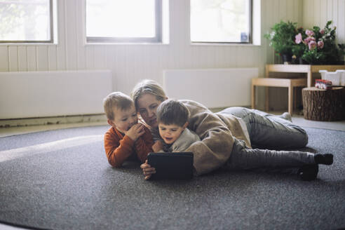 Female teacher watching digital tablet with boys while lying down on carpet at preschool - MASF43140