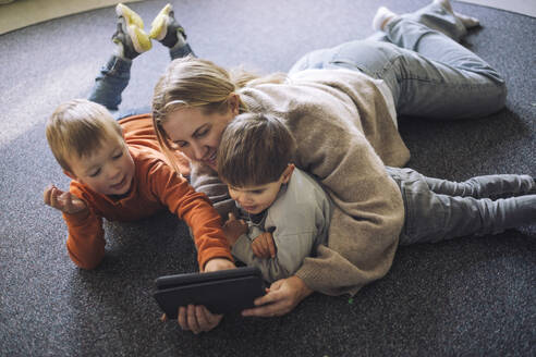 High angle view of kids watching digital tablet with female teacher lying down on carpet at preschool - MASF43141