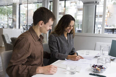 Man looking at woman smiling while signing agreement at desk in real estate office - MASF43152
