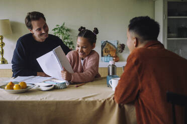 Happy daughter showing notebook to gay father at dining table in home - MASF43163