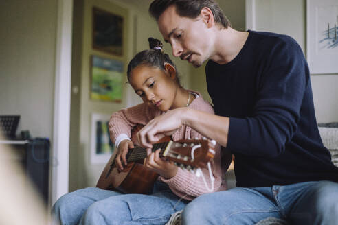 Father assisting daughter playing guitar while sitting on bed at home - MASF43166