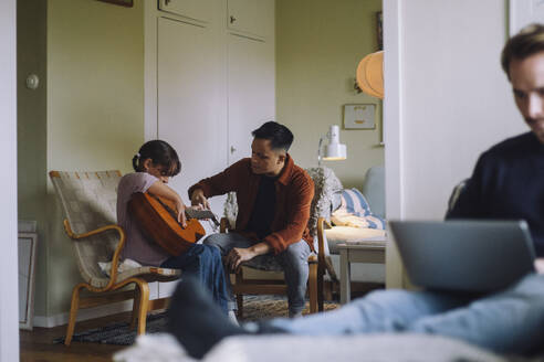 Father teaching daughter to play guitar while sitting on chair at home - MASF43168
