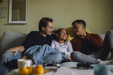Smiling gay men spending leisure time with daughter while sitting on sofa at home - MASF43177