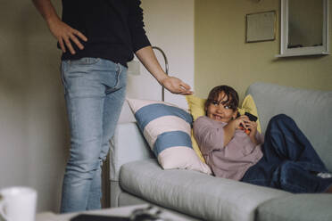 Midsection of father asking smart phone from daughter sitting on sofa at home - MASF43182