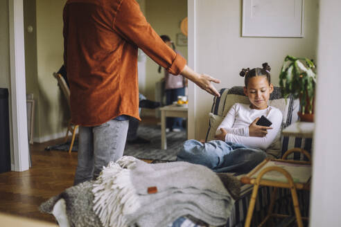 Midsection of father asking smart phone from daughter sitting on bed at home - MASF43185