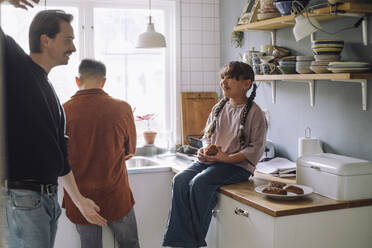 Father playing with daughter holding bread while sitting on kitchen counter at home - MASF43199