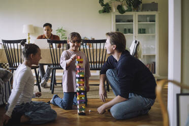 Smiling girl stacking Jenga blocks while kneeling on floor by father and sister at home - MASF43205