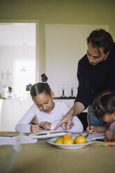 Father assisting to daughters while doing homework sitting at dining table - MASF43213