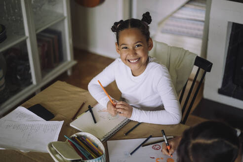 High angle portrait of happy girl sitting at table holding colored pencil - MASF43216