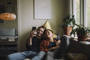 Smiling gay couple sharing smart phone while sitting near window at home - MASF43218