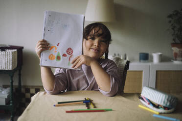 Portrait of smiling girl showing drawing on notebook while sitting at home - MASF43229