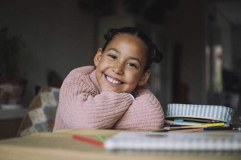 Portrait of happy girl resting head on arms while sitting on table with colored pencils at home - MASF43236