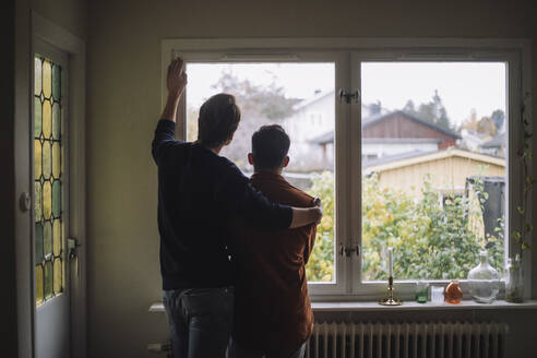 Rear view of man with arm around gay couple looking through window at home - MASF43250