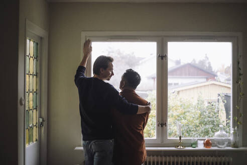 Rear view of romantic gay couple standing near window at home - MASF43251