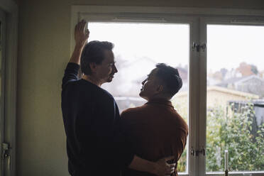 Rear view of multiracial gay couple talking while standing near window at home - MASF43252