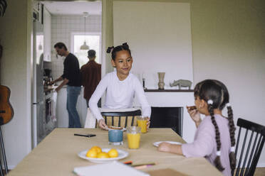 Smiling girl looking at sister while setting table for breakfast at home - MASF43262