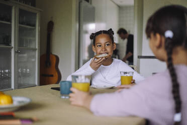 Girl eating bread slice while sitting at dining table with sister in home - MASF43263