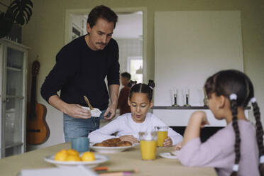 Father serving cheese spread to daughter having breakfast at dining table in home - MASF43264