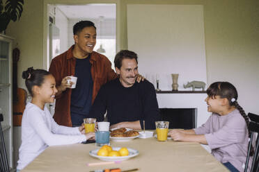 Happy gay couple having fun while having breakfast with daughters at dining table in home - MASF43266