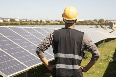 Rear view of male engineer with arms akimbo looking at solar panels in field - MASF43270