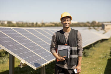 Portrait of smiling male engineer holding documents and digital tablet while standing near solar panels at power station - MASF43273