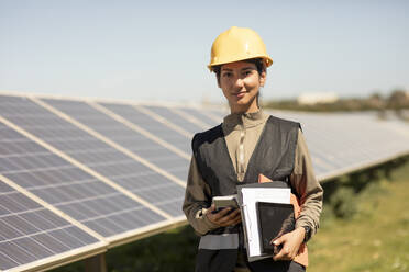 Portrait of smiling female maintenance engineer holding smart phone and documents while standing near solar panels in fi - MASF43274