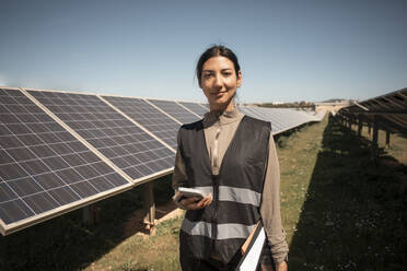 Portrait of smiling female maintenance engineer holding smart phone while standing near solar panels in field - MASF43275