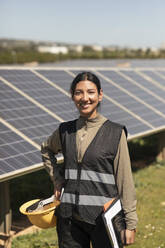 Portrait of confident female engineer standing with hand on hip near solar panels in field - MASF43276