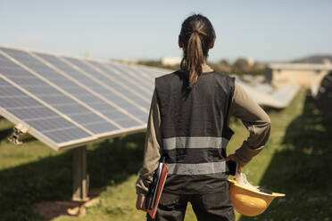 Rear view of female engineer standing with hand on hip while looking at solar panels in field - MASF43277