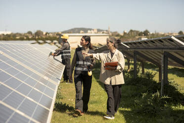 Female entrepreneur examining solar panels while walking with engineer at power station - MASF43286