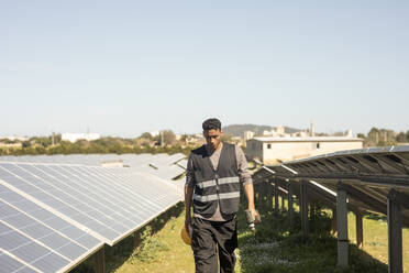 Young male maintenance engineer holding drill while walking amidst solar panels at power station - MASF43291