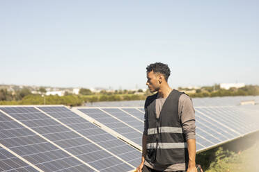 Young male engineer wearing reflective jacket while looking at solar panels at power station - MASF43292