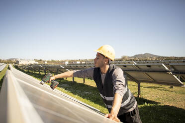 Young male engineer drilling while working near solar panels at power station - MASF43297