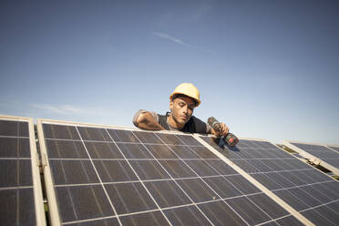 Young male engineer holding drill while examining solar panels at power station - MASF43299