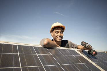 Portrait of smiling male engineer holding drill while leaning on solar panels at power station - MASF43300