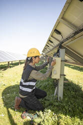 Side view of female engineer repairing fuse box while kneeling near solar panels at power station - MASF43306