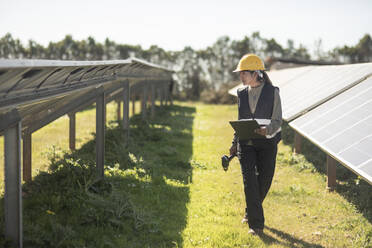 Female maintenance engineer examining solar panels while walking at power station - MASF43307