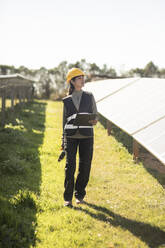 Female engineer looking away while walking amidst solar panels at power station - MASF43308