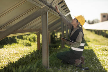 Side view of female engineer examining solar panels while kneeling at power station - MASF43309