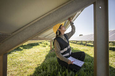 Female maintenance engineer working while kneeling under solar panels at power station - MASF43311