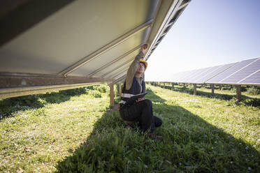 Female engineer repairing solar panels while kneeling on grass at power station - MASF43312