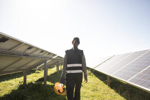 Rear view of female engineer holding hard hat while walking amidst solar panels at power station - MASF43317