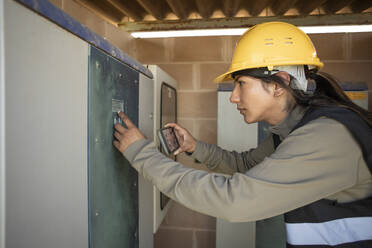 Side view of female engineer programming fuse box through smart phone at power station - MASF43320