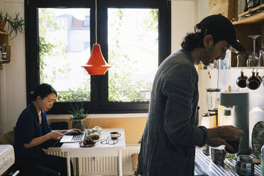 Male architect preparing coffee while female colleague using smart phone in home office - MASF43329