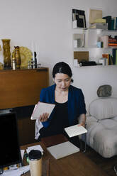 Mature female architect holding diary while standing at desk in home office - MASF43339